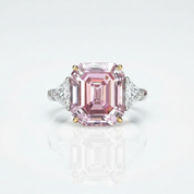 An emerald-cut pink diamond ring with triangular diamond side stones in a silver and gold setting, centered on a clean white background with a soft reflection below.