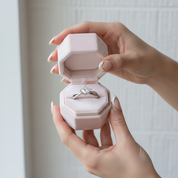 Hand holding a pink jewelry box with a ring inside against a neutral background