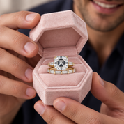 A close-up lifestyle shot of a person holding an open dusty pink velvet ring box, showcasing a yellow gold engagement ring with a large emerald-cut diamond and a matching diamond band.