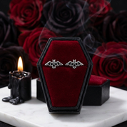 A pair of silver bat-shaped stud earrings displayed on a black coffin-shaped velvet box with a deep red interior, set on a white marble surface with dark red roses and a lit black candle in the background.