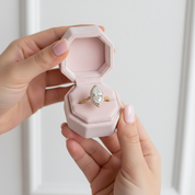 A close-up of a person's hands holding an open light pink velvet ring box, displaying a gold ring with a marquise-cut clear gemstone.