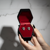Black hexagonal jewelry box with red interior holding silver skull earrings, held by a hand on a marble surface.