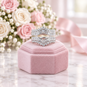 A pair of silver-toned chevron ring sets with baguette-cut clear stones, displayed on a pink velvet ring box with a bouquet of pink and white roses in the background.