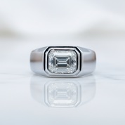 A silver-toned signet ring featuring a large emerald-cut clear gemstone, displayed on a reflective white surface against a soft, out-of-focus light gray background.