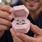 A silver ring with black and white diamond accents is displayed inside an open pink velvet hexagonal ring box, held by a person whose hands and lower face are visible.
