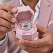 A close-up of a person holding an open pink velvet ring box containing two silver ring sets with clear stones. The person is wearing a light pink blazer and white dress shirt.