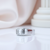 A silver-toned signet ring with a large rectangular clear gemstone, resting on a reflective white surface with a soft-focus background.