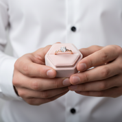 Person holding a pink hexagonal jewelry box with a ring inside, wearing a white shirt.