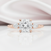 A rose gold engagement ring featuring a large cushion-cut diamond center stone and two smaller round diamonds on the shoulders, set against a soft, light pink and white background.