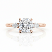 A rose gold engagement ring featuring a large cushion-cut diamond center stone and two smaller round diamonds on the shoulders, set against a clean white background with a soft reflection.