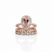 A rose gold ring set featuring a pear-cut pink gemstone surrounded by a halo of small clear stones, with a matching rose gold band set with round clear stones, displayed on a clean white background with a soft reflection.