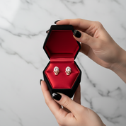 Skull-shaped earrings in a hexagonal black box with red interior held by a hand against a marble background.