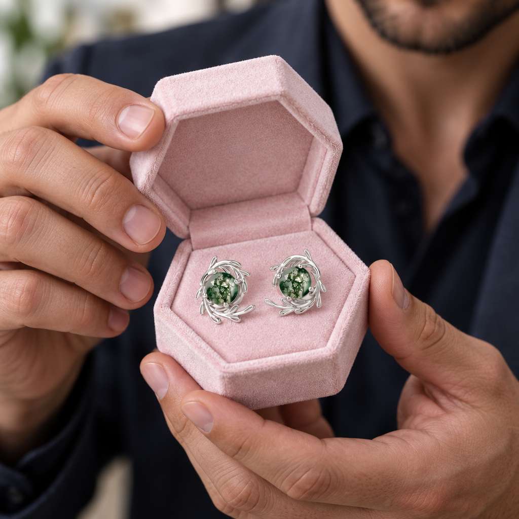 A close-up of a person holding an open pink jewelry box with a pair of silver-toned earrings featuring green gemstone centers.