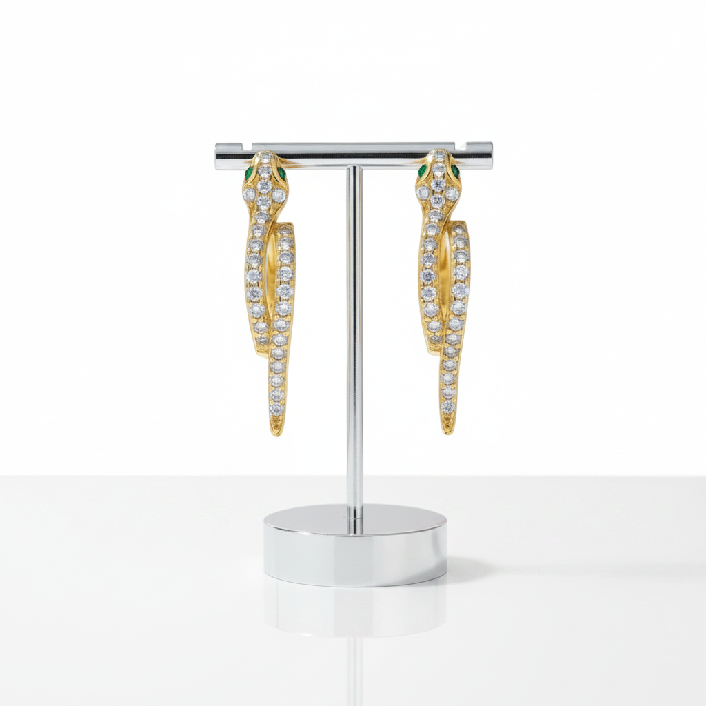 A pair of gold snake-shaped hoop earrings with green enamel details and rows of clear pavé stones, displayed on a polished silver stand against a clean white background.
