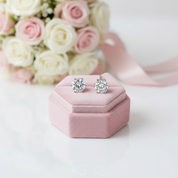 A pair of round-cut diamond stud earrings in a silver setting, displayed on a small pink velvet jewelry box with a soft-focus bouquet of pink and white roses in the background.