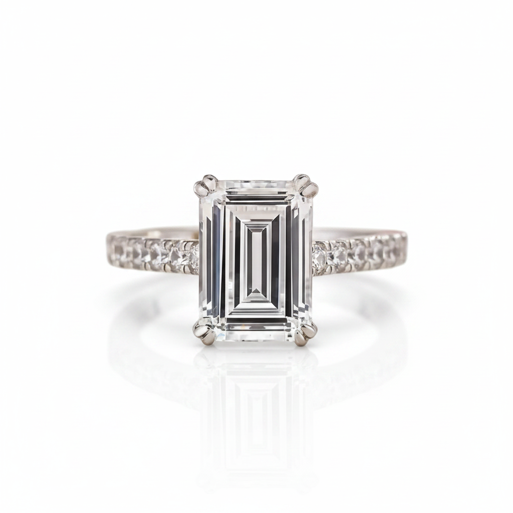 An emerald-cut diamond engagement ring with a four-prong setting and a diamond-paved band, shown on a clean white background with a soft reflection.