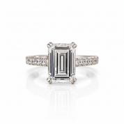 An emerald-cut diamond engagement ring with a four-prong setting and a diamond-paved band, shown on a clean white background with a soft reflection.