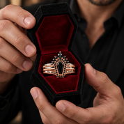 A close-up of a person holding an open black velvet ring box with a gothic coffin ring inside. The ring features a black coffin-shaped stone set in rose gold with small black gemstones and the words 'I WILL DEATH' and 'I MUST LIVE' engraved on the bands.