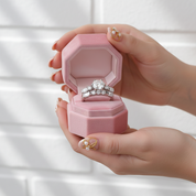 Hand holding a pink jewelry box with a diamond ring inside against a neutral background