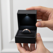 Hand holding a black jewelry box with a silver ring inside against a white background
