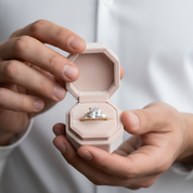 Engagement ring in a pink box held by two hands against a neutral background