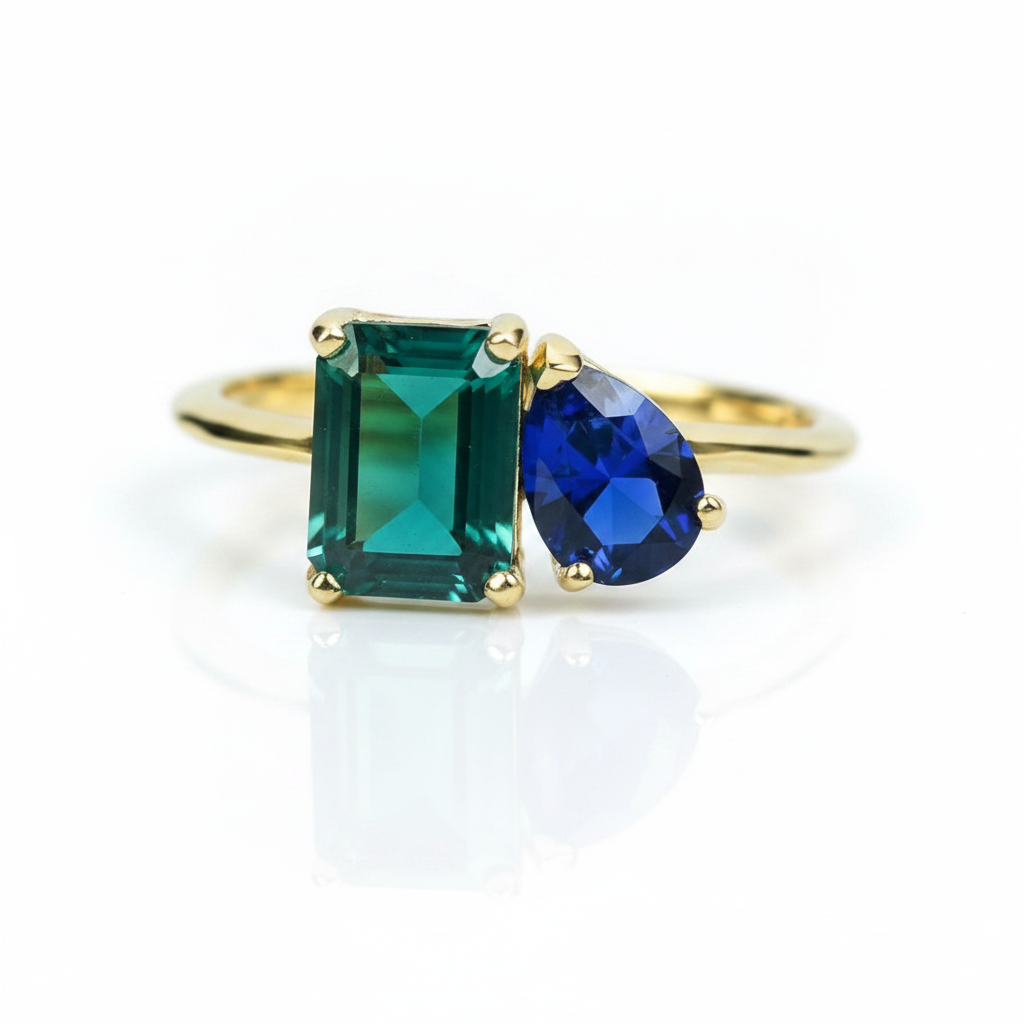 A gold ring featuring a rectangular emerald-cut green gemstone and a pear-cut deep blue gemstone, set on a clean white background with a soft reflection.