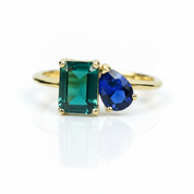 A gold ring featuring a rectangular emerald-cut green gemstone and a pear-cut deep blue gemstone, set on a clean white background with a soft reflection.