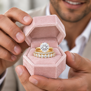A close-up lifestyle shot of a man holding an open pink velvet ring box. Inside, a gold ring with a large round-cut diamond and a matching diamond band are displayed.