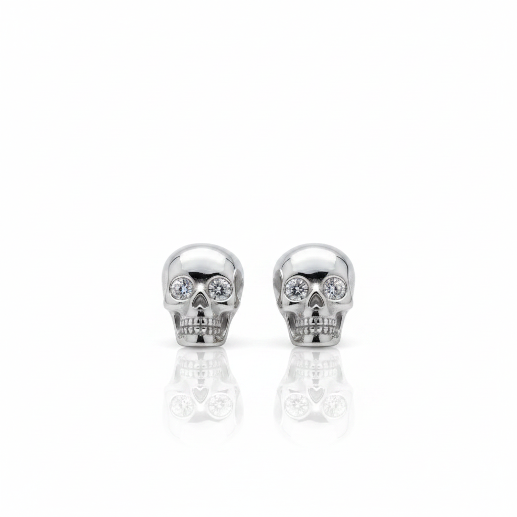 A pair of silver skull stud earrings with clear crystal eyes, displayed on a clean white background with a soft reflection below.